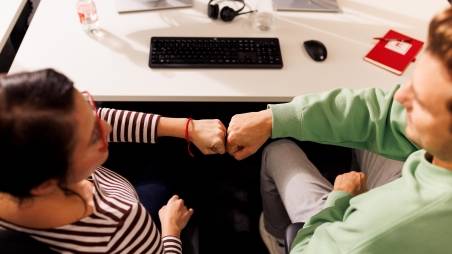 Kollege und Kollegin sitzen gemeinsam im Kaufland-Büro am Schreibtisch mit schwarzer Tastatur. Ihre zur Faust geballten Hände berühren sich zum Gruß.